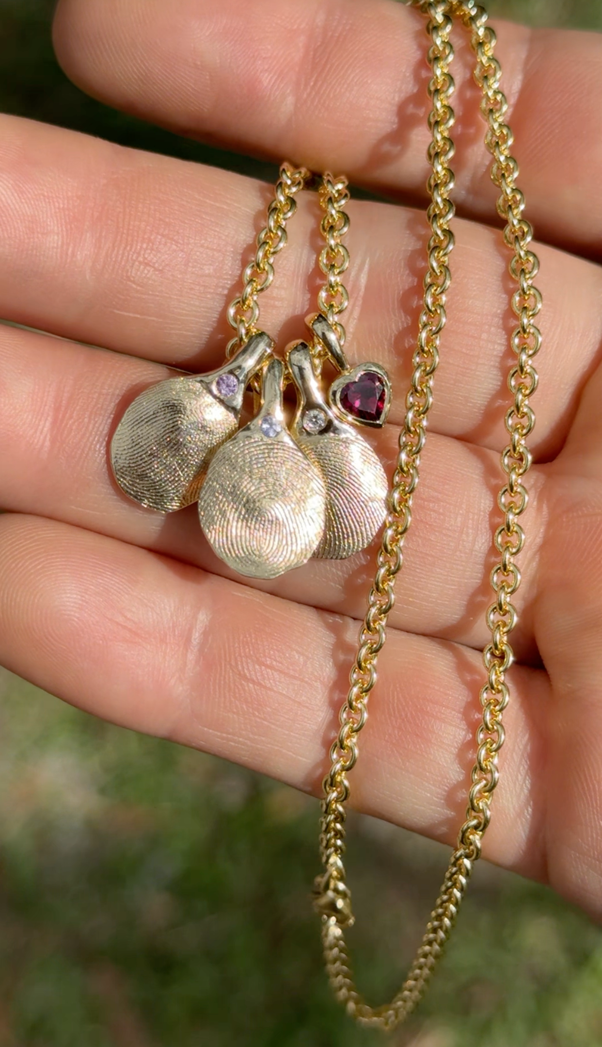 A gold chain necklace with three textured pendant charms, including a small purple heart and two teardrop-shaped pendants, held in a person's hand against a blurred green outdoor background.